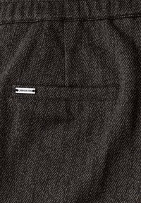 Detailní záběr na látku v šedém a černém herringbone vzoru s kapsou na slitinu, na které je malá kovová značka s nápisem "Street One."