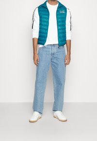 Zielonkawa kamizelka puchowa na białej koszuli z długim rękawem, zestawiona z jasnoniebieskimi dżinsami i białymi tenisówkami, prezentująca swobodny, sportowy wygląd.