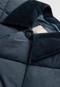 Jachetă puffer de culoare bleumarin, cu guler din catifea, închidere cu nasturi și textură matlasată. Materialul este neted, cu un finisaj ușor lucios.