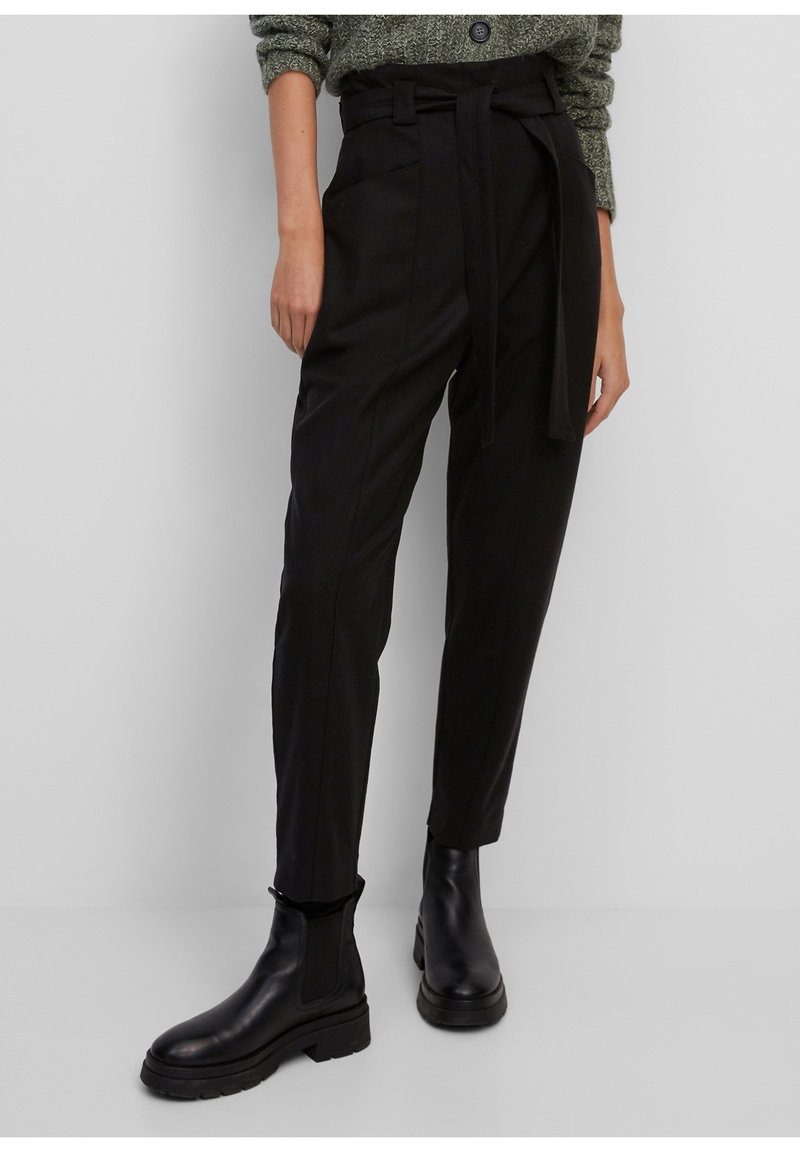 Pantalons noirs taille haute avec une ceinture nouée, poches latérales et une jambe fuselée, associés à des bottines en cuir noir.