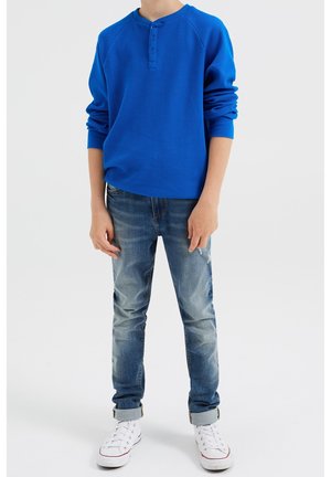 Maglietta blu a maniche lunghe con collo henley, abbinata a jeans in denim strappati e sneakers alte bianche.
