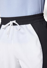 Pantalones cortos deportivos blancos con acentos laterales negros, una cinturilla elástica y un cordón blanco. Tejido texturizado con un patrón de cuadrícula.