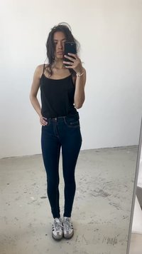 Top preto sem mangas combinado com jeans skinny azul-escuro e ténis cinzentos. O conjunto apresenta um design simples com uma silhueta ajustada.