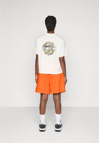 Hvit kortermet skjorte med et sirkulært grafisk trykk på baksiden; kombinert med oransje shorts. Sokker og joggesko kompletterer antrekket.