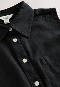 Schwarzes Leinenhemd mit klassischem Kragen, acht weißen Knöpfen, Tasche auf der linken Seite und Spitzenverzierung nahe dem Kragen. Glatte Textur.