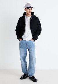 Chaqueta bomber negra, camiseta blanca, jeans azul claro, zapatos negros y gorra gris con visera oscura. Modelo de pie con las manos en los bolsillos.