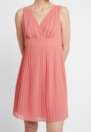Mouwloze koraalroze jurk met V-hals en brede tailleband, met een geplooide rok die boven de knieën valt, gedragen door een staand persoon.