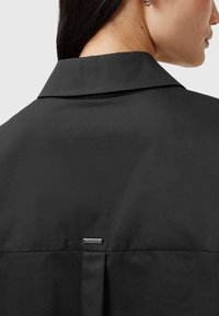 Camicia nera con colletto caratterizzata da una texture liscia, una leggera lucentezza e un piccolo etichetta metallica sul retro. Design strutturato con dettagli plissettati.