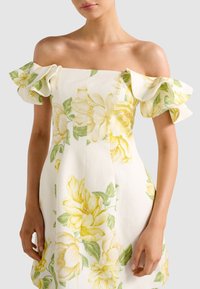 Γυναίκα με λευκό φόρεμα έξω από τους ώμους με κίτρινο floral μοτίβο και φουσκωτά μανίκια, στέκεται μπροστά σε απλό φωτεινό φόντο.