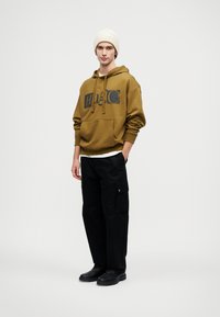 Brauner Hoodie mit einem Vorderdesign, ausgestattet mit einer Kängurutasche, kombiniert mit schwarzen Cargo-Hosen mit Seitenfächern und schwarzen Schuhen.