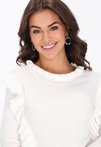 Biały sweter z prążkowanej tkaniny z falbanami na ramionach. Tekstura jest miękka, a projekt charakteryzuje się okrągłym dekoldem.