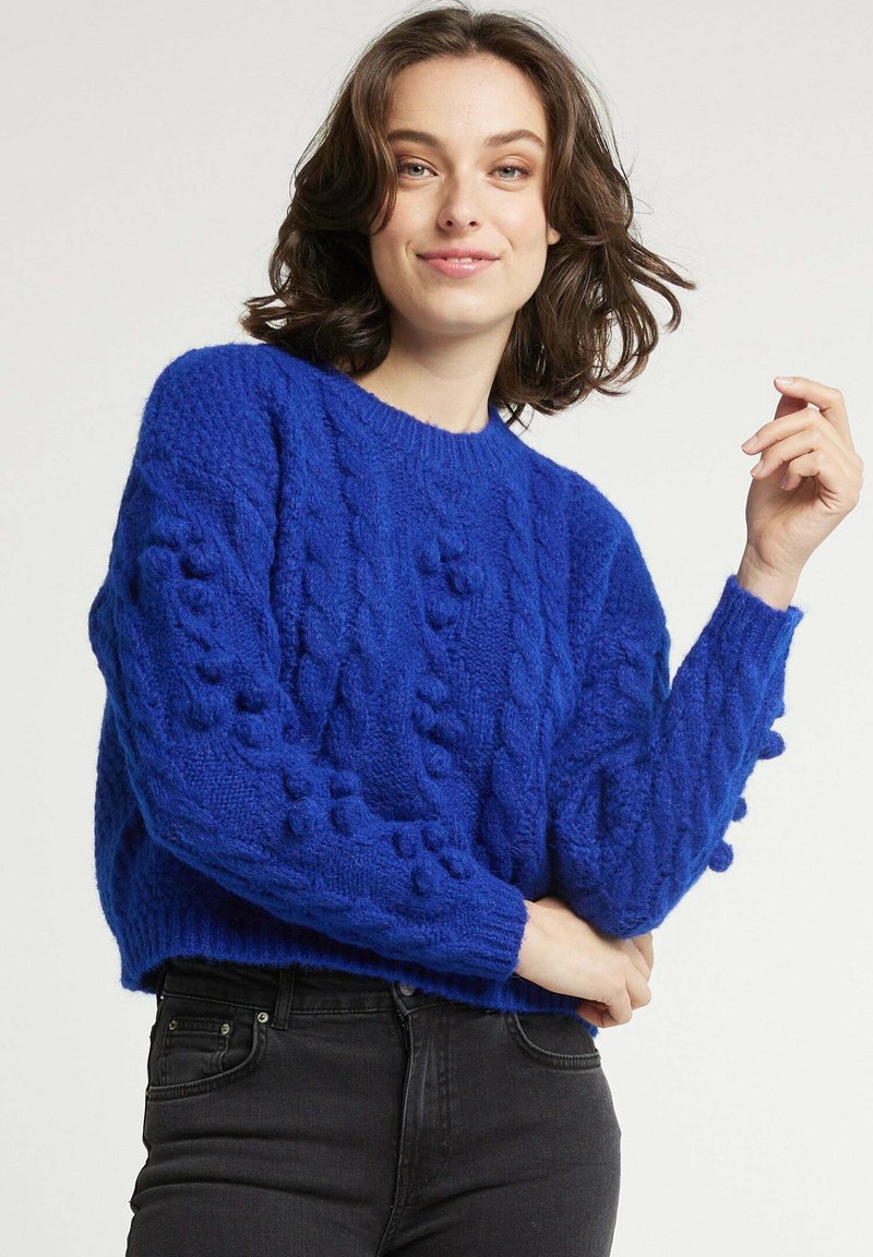 Pull en maille torsadée bleu avec des détails en relief, col rond et manches longues, associé à un pantalon noir. Matière en tricot épaisse et confortable.