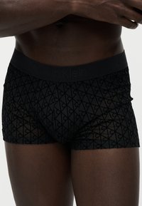 Torse masculin portant un boxer noir à motifs avec une ceinture Calvin Klein discrète, mains croisées près de la poitrine.