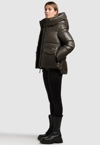 Tamsiai ruda pūkinė striukė su aukštu apykakle, dviem priekinėmis kišenėmis, blizgia medžiaga ir prigludusia forma, derinama su juodais ilgaauliais batais.