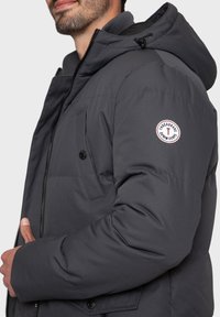 Dunkelgraue Steppjacke mit Kapuze, ausgestattet mit einem Frontreißverschluss, Druckknopftaschen und einem runden Logoaufnäher am linken Ärmel. Glatte Textur.