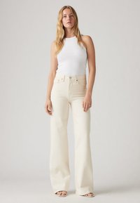 Calças de ganga creme, de cintura alta e perna larga, feitas de denim, com fecho de botão e detalhes subtis de costura laranja. Combinadas com um top de alças branco.