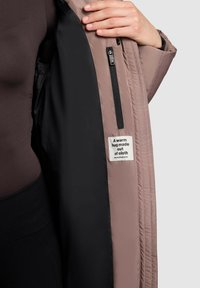 Puffer jacket with a black interior and a soft mauve exterior. Features a zippered pocket and a label stating, "A warm hug made out of cloth."