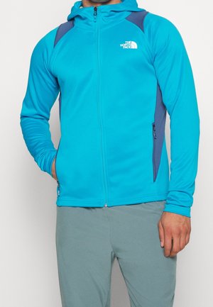 Homme portant une veste à capuche zippée bleu vif avec des panneaux aux épaules et sur les côtés bleu foncé, logo blanc « The North Face » sur la poitrine.