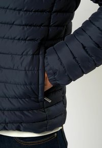 Navy puffersjakke med kviltet tekstur, elastiske mansjetter og sidelommer. Har en subtil metallisk merkelapp nær kanten.