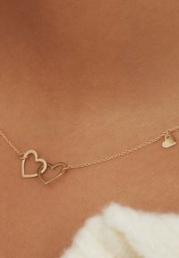 Goldene Halskette mit zwei ineinandergreifenden Herzanhängern und einem kleinen hängenden Herzdetail, vor einem Hautton-Hintergrund.