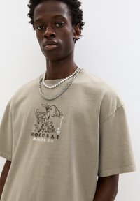 Khaki oversized tričko s černým grafickým potiskem znázorňujícím květiny a text, s kulatým výstřihem a krátkými rukávy, nošeno s vrstvenými náhrdelníky.