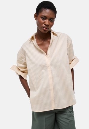 Femme aux cheveux courts portant une chemise ample à rayures beige clair avec des manches retroussées et un pantalon vert, debout les mains dans les poches.