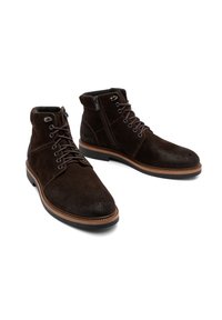 Brązowe zamszowe botki za kostkę z okrągłymi oczkami, brązowymi sznurówkami i bocznym zamkiem. Posiadają beżowy akcent wokół podeszwy oraz teksturowany wierzch.