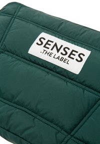 Gepolsterte grüne Tasche mit gestepptem Muster. Verfügt über ein weißes Etikett mit schwarzem Text "SENSES .THE LABEL." Genähte Akzente werten das Design auf.