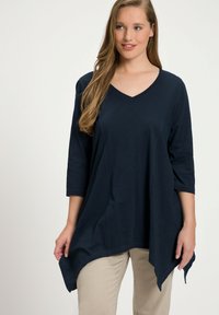 Top blu navy a V con maniche 3/4, caratterizzato da un orlo asimmetrico e una consistenza morbida e leggera. Abbinato a pantaloni beige.