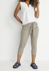 Lehká bílá bezramínková blůza s výstřihem do V a volánkovým lemem, kombinovaná s světle béžovými zkrácenými kalhotami s vázáním v pase a modrými sandály.