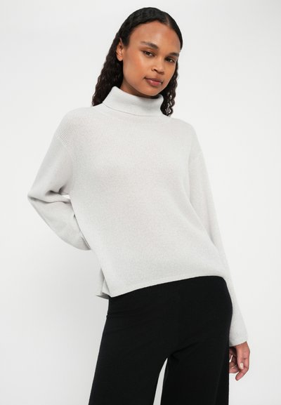 Pull en maille côtelée gris clair à col roulé, avec des manches longues et des coutures d'épaule tombantes, associé à un pantalon noir. Texture douce et coupe décontractée.
