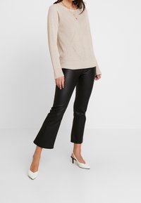 Kobieta nosi beżowy sweter z dzianiny, czarne skórzane spodnie cropped i białe buty na obcasie typu slingback, stojąc na tle jasnego, gładkiego tła.