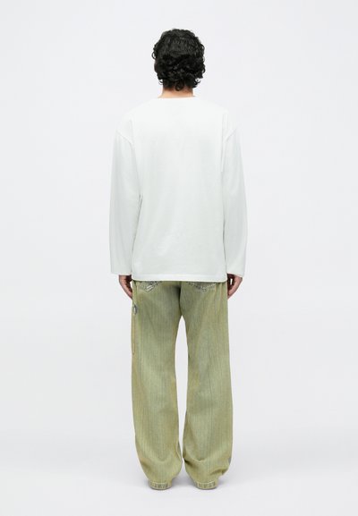 Vīrietis ar cirtainiem matiem stāv ar muguru pret skatītāju, valkājot baltu garu krekla piedurkni un vaļīgas, gaiši zaļas džinsa bikses.