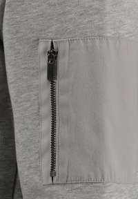 Felpa grigia con una sezione superiore testurizzata e una sezione inferiore liscia, dotata di una tasca con zip sul braccio sinistro.