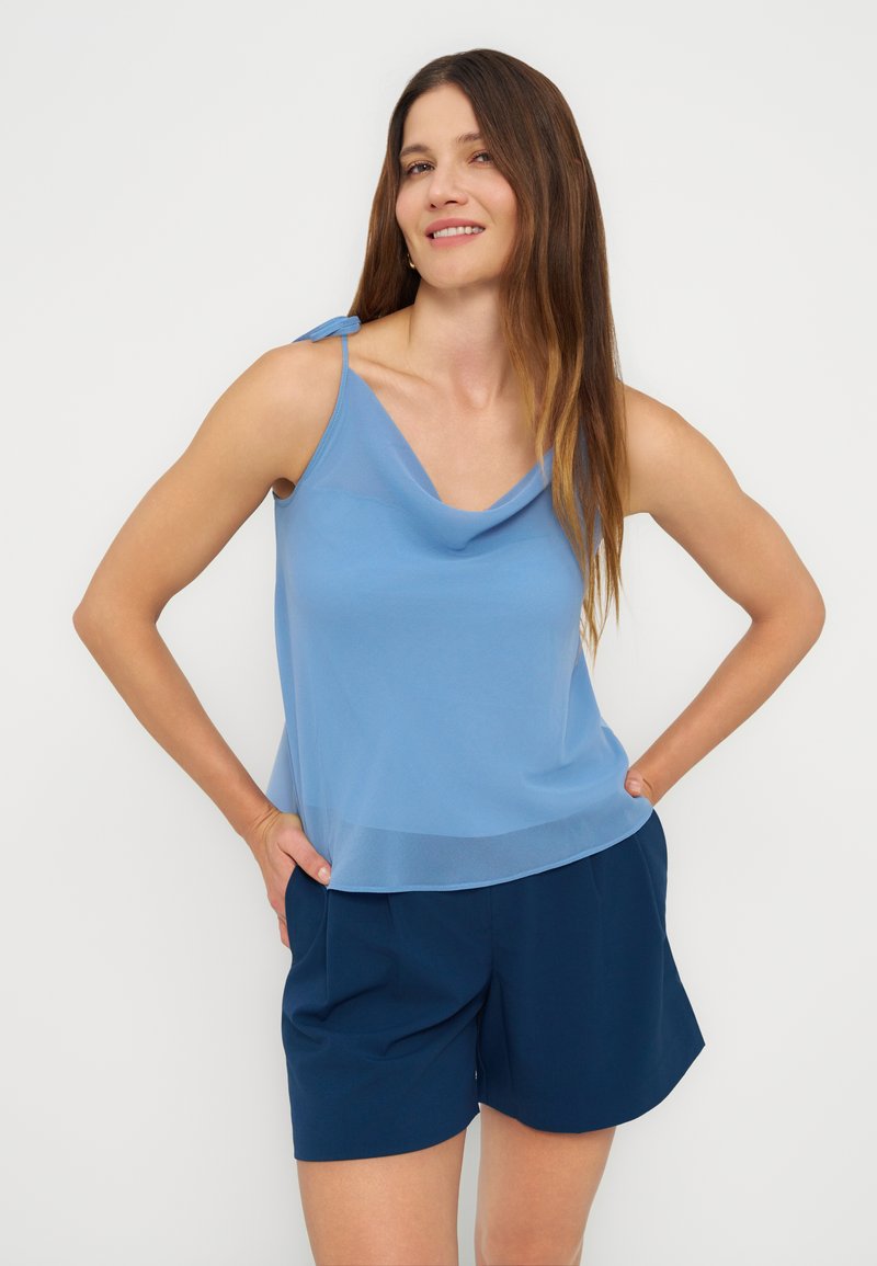 Femme aux longs cheveux bruns portant un haut sans manches bleu clair et un short bleu foncé, souriante avec les mains sur les hanches sur fond blanc.