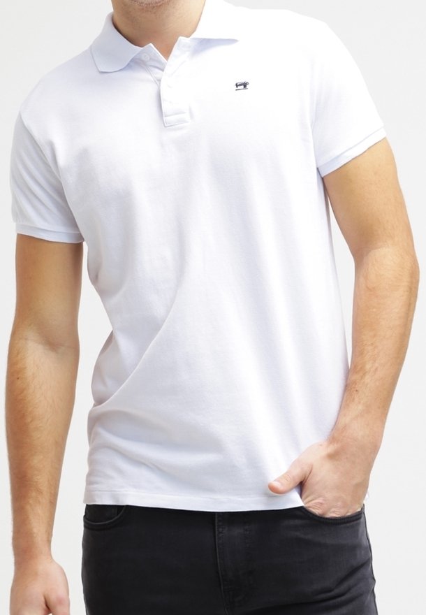 Homme portant un polo à manches courtes blanc uni avec un petit logo noir sur la poitrine, la main dans la poche, et un pantalon noir.