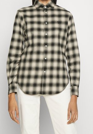 Kvinde iført sort- og hvidternet langærmet skjorte med knapper og hvide bukser, stående foran en ensfarvet baggrund.