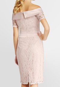  pitsimekko pehmeässä vaaleanpunaisessa sävyssä, jossa on olkapäiden päältä avautuva satiinisomiste, muotoon ommeltu siluetti, takakuvioinen vetoketju ja pitsiyksityiskohtia, joissa on halkio helmoissa.