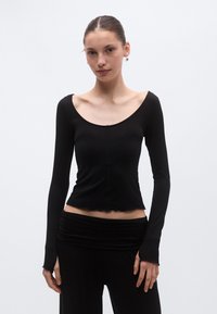 Μαύρο μπλουζάκι μακρυμάνικο με στρογγυλεμένο ντεκολτέ, εφαρμοστή γραμμή, ριμπ υφή και κυματιστό τελείωμα, φορεμένο με συνδυαστικό μαύρο παντελόνι.