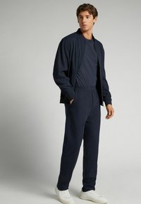 Tmavě modrý teplákový komplet z měkké tkaniny, obsahující bomber bundu na zip a odpovídající kalhoty s zúženým střihem, doplněné o bílé tenisky.