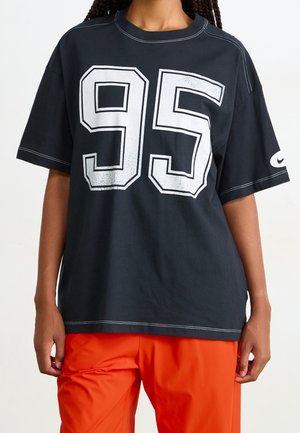 Osoba nosi czarną oversize koszulkę z dużą białą cyfrą "95" na przodzie i pomarańczowe spodnie, pokazując logo Nike na rękawie.