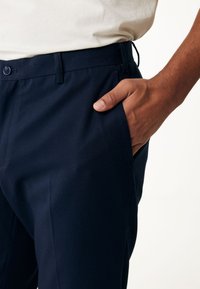 Marinblå skräddarsydda byxor med en slät yta, sidofickor och knäppning med knapp. Löst sittande design med bälteshällor och rena, raka linjer.