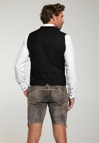 Homme de dos portant une chemise blanche, un gilet noir avec une sangle ajustable, et un short en cuir brun foncé avec des lacets sur les côtés et des coutures décoratives.