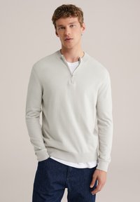 Pull-over gris clair à fermeture éclair en tissu doux tricoté, avec un col rond, des manches longues et une coupe décontractée. Porté par-dessus une chemise blanche.