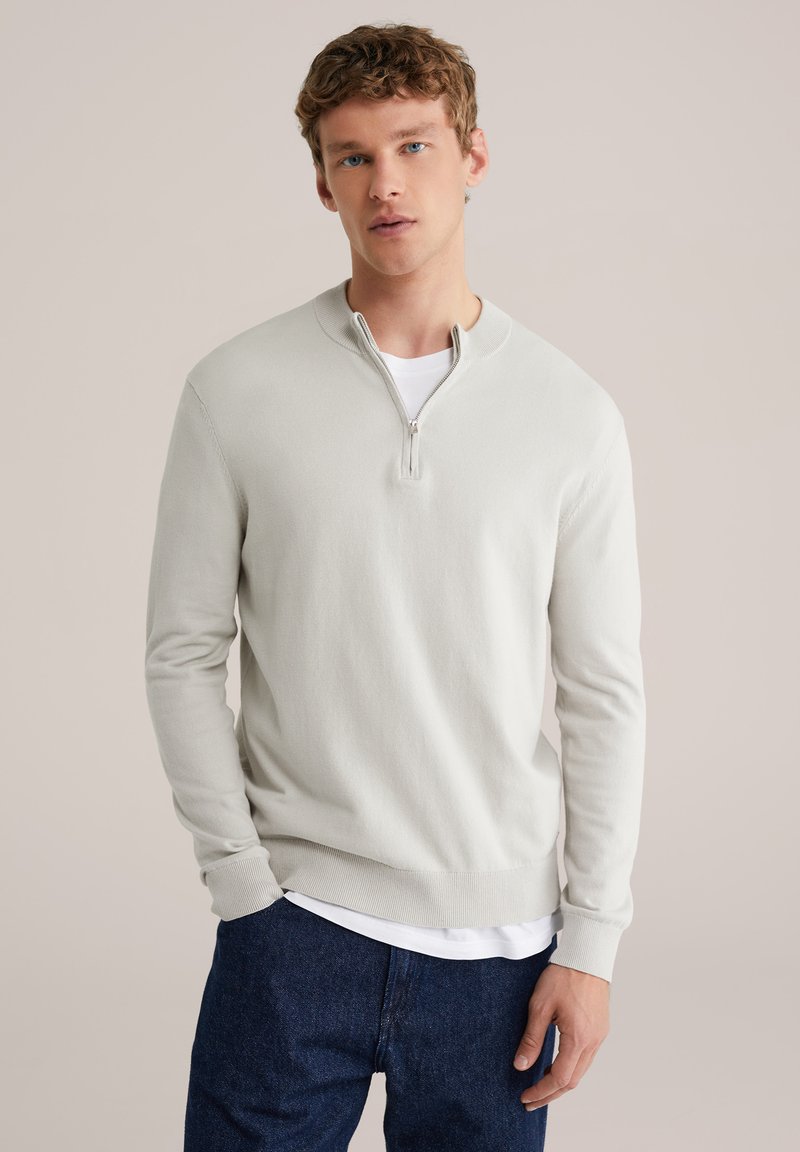 Pull-over gris clair à fermeture éclair en tissu doux tricoté, avec un col rond, des manches longues et une coupe décontractée. Porté par-dessus une chemise blanche.