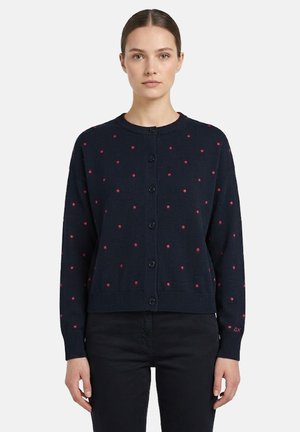 Donna che indossa un cardigan blu navy con bottoni e piccoli pois rossi, abbinato a pantaloni neri, in piedi davanti a uno sfondo neutro.