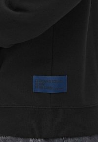 Etichetta in tessuto blu con testo nero "Dedicate alla perfezione del denim" cucita sulla parte bassa di una felpa nera sopra a un paio di jeans blu.