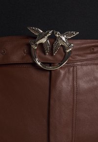 Boucle de ceinture en argent avec deux oiseaux faisant face l'un à l'autre sur une ceinture en cuir marron portée sur un tissu foncé.