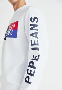 Sudadera blanca con un logotipo rectangular rojo y azul en el frente. La manga izquierda tiene un texto en negrita negro: "PEPE JEANS". Textura de tela suave.
