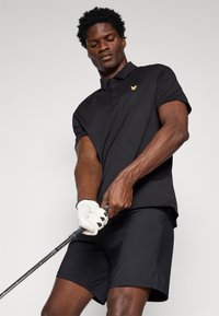 Melns polo krekls ar īsu apkakli un zelta emblēmu, apvienots ar melnām šortiem. Persona tur golfu nūju, valkājot baltu cimdu.